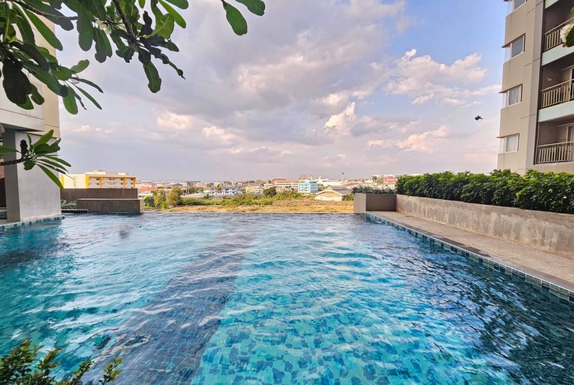 Pool edged by modern buildings, blue-tiled water, and a leafy foreground branch with a distant city skyline under a partly cloudy sky.