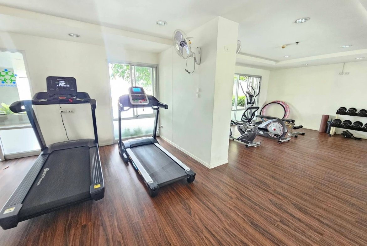 Gym cardio area with two treadmills near large windows and wood floor. A cross between cardio machines and weights visible in the background.