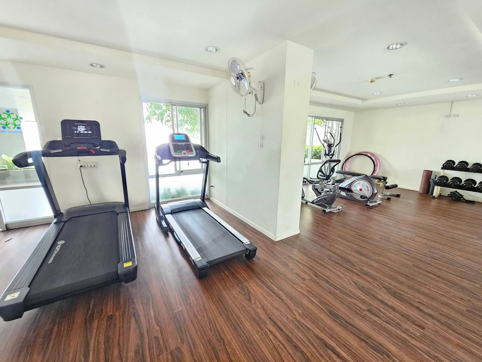 Lumpini_Condo_Town_North_Pattaya_Sukhumvit 6 Gym cardio area with two treadmills near large windows and wood floor. A cross between cardio machines and weights visible in the background.
