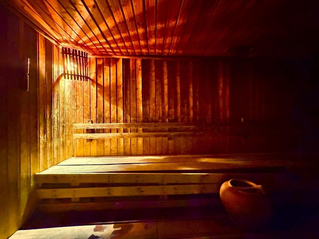 Wood-paneled sauna with tiered benches, warm amber lighting, and a ceramic pot on the floor at the corner.