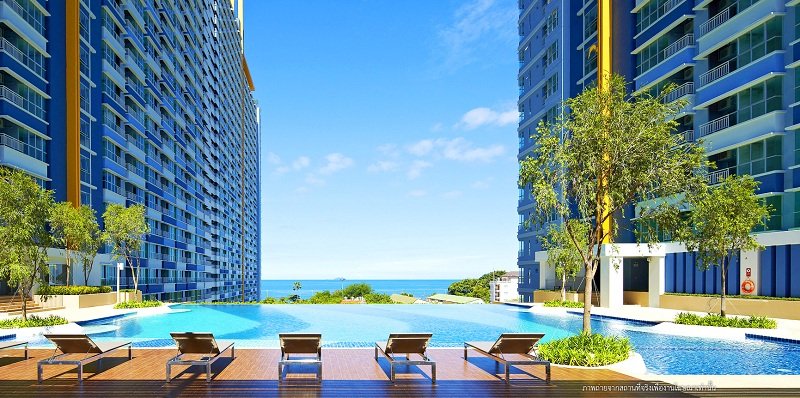 Resort pool area between two tall blue apartment buildings, lounge chairs facing a sunny ocean view.