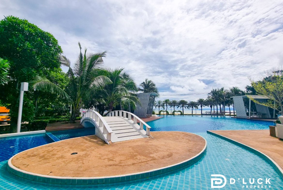 Resort pool with a curved sandy island and white footbridge, surrounded by palm trees under a cloudy sky; D'Luck Property logo in corner.