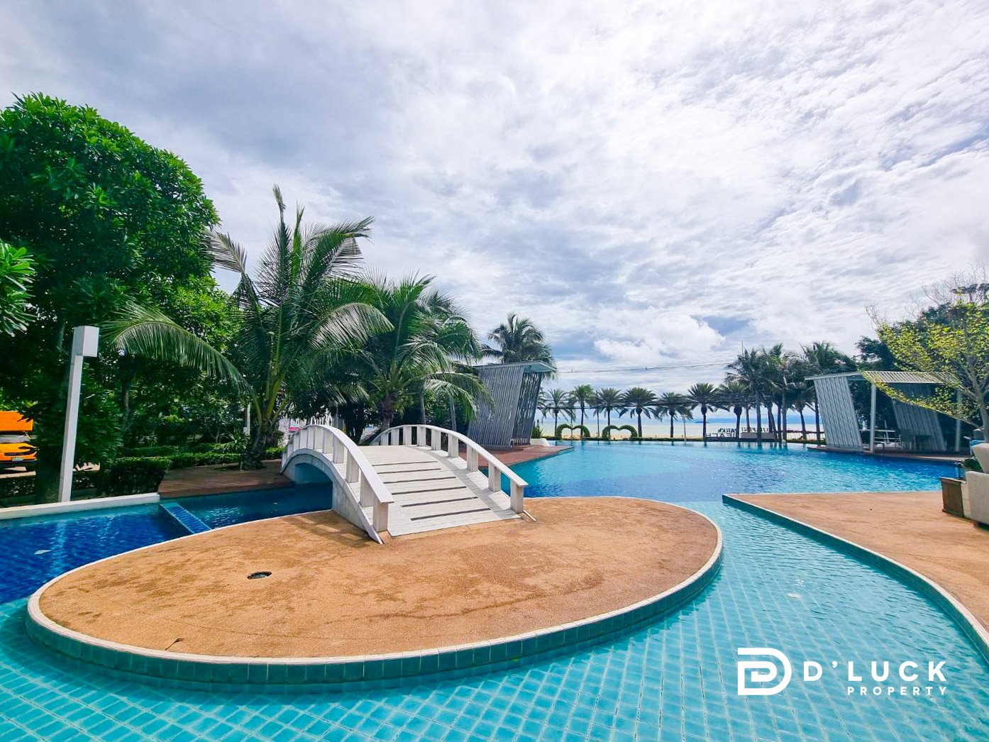 Lumpini_Park_Beach_Jomtien_Pattaya 2 Resort pool with a curved sandy island and white footbridge, surrounded by palm trees under a cloudy sky; D'Luck Property logo in corner.