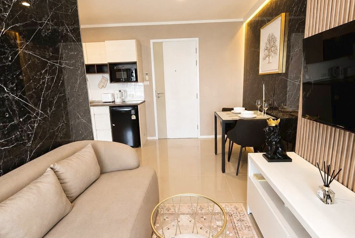 Open-plan living area with a beige sofa, black marble accent wall, glass coffee table, white TV console, and a two-seat dining table in the background; kitchen seen beyond the door.