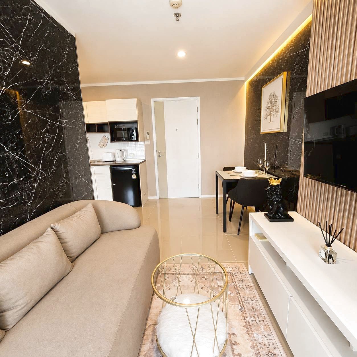 Lumpini_Park_Beach_Sale_Dlc650 Open-plan living area with a beige sofa, black marble accent wall, glass coffee table, white TV console, and a two-seat dining table in the background; kitchen seen beyond the door.