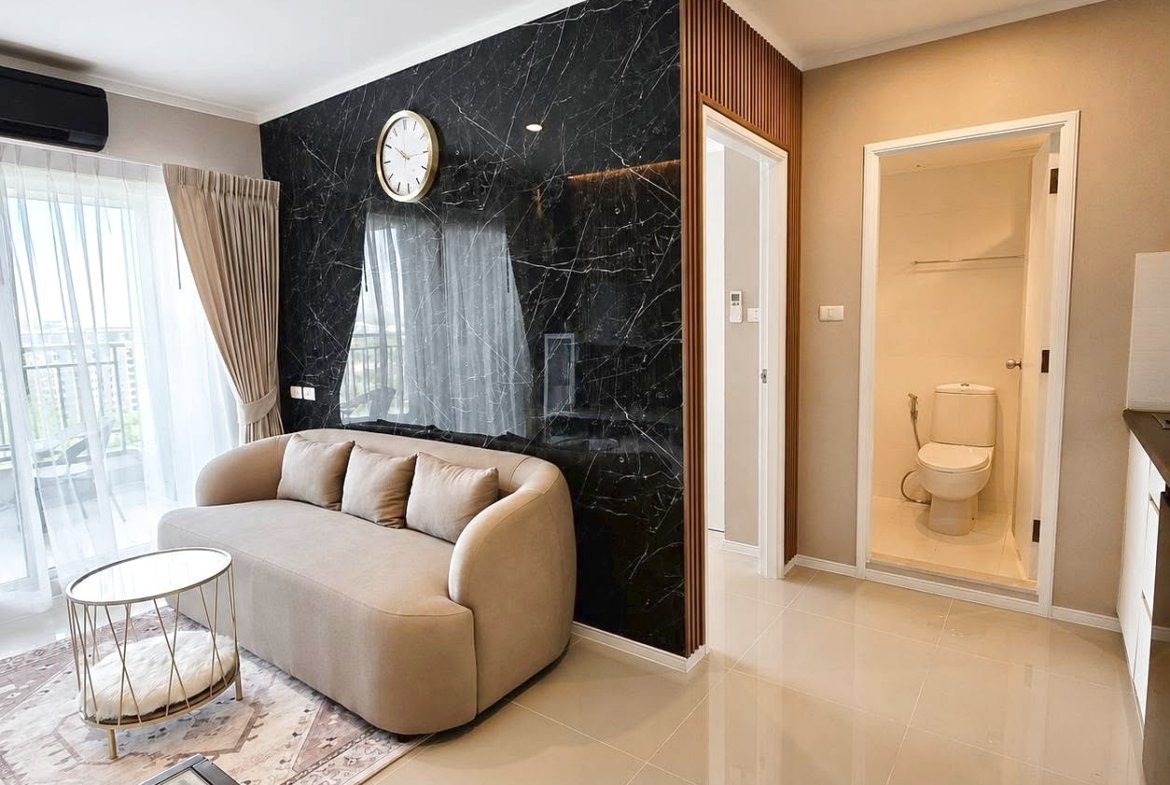 Living room with a curved beige sofa, light cushions, and a black marble feature wall with a wall clock. Large glass doors lead to a balcony with sheer curtains.