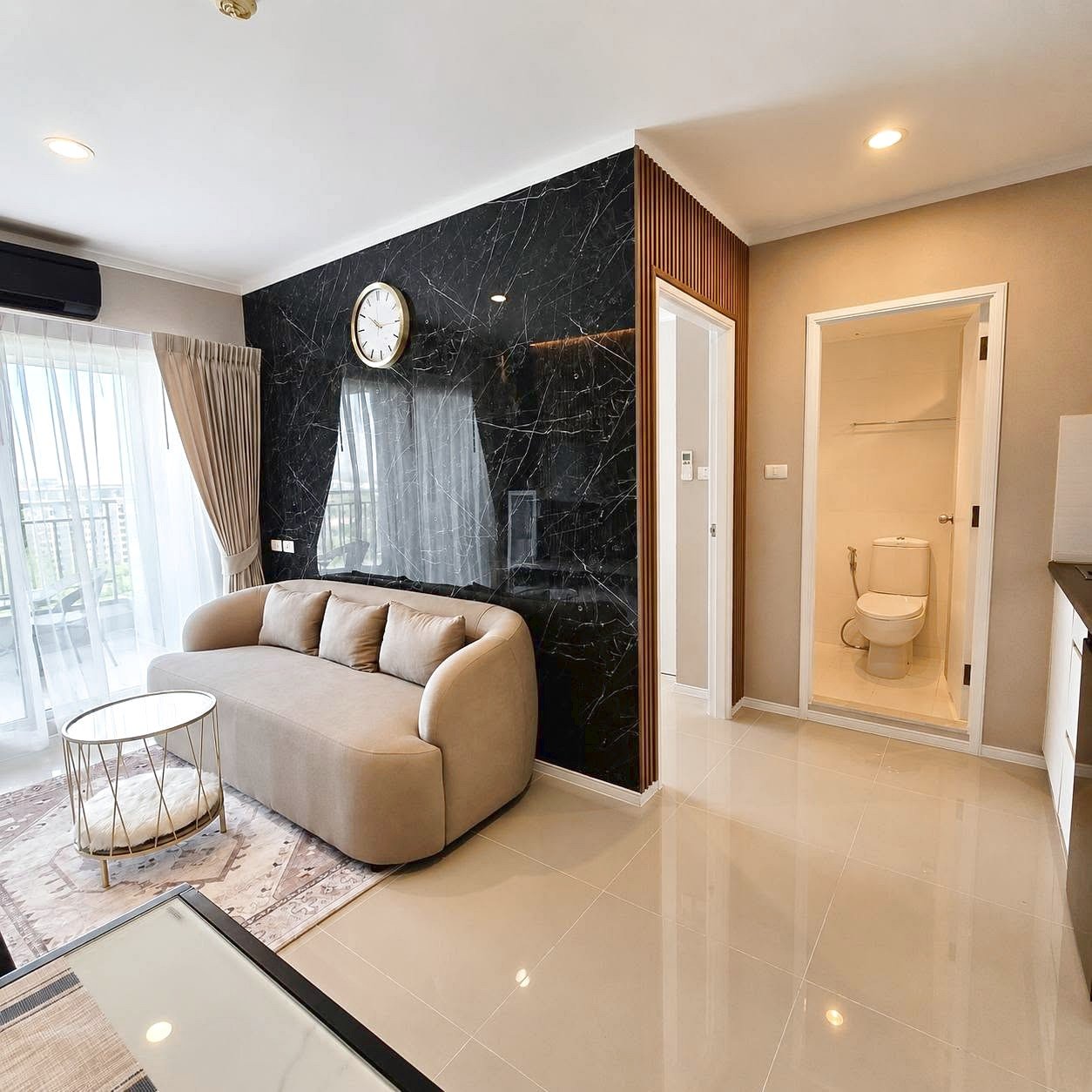Lumpini_Park_Beach_Sale_Dlc650_12 Living room with a curved beige sofa, light cushions, and a black marble feature wall with a wall clock. Large glass doors lead to a balcony with sheer curtains.