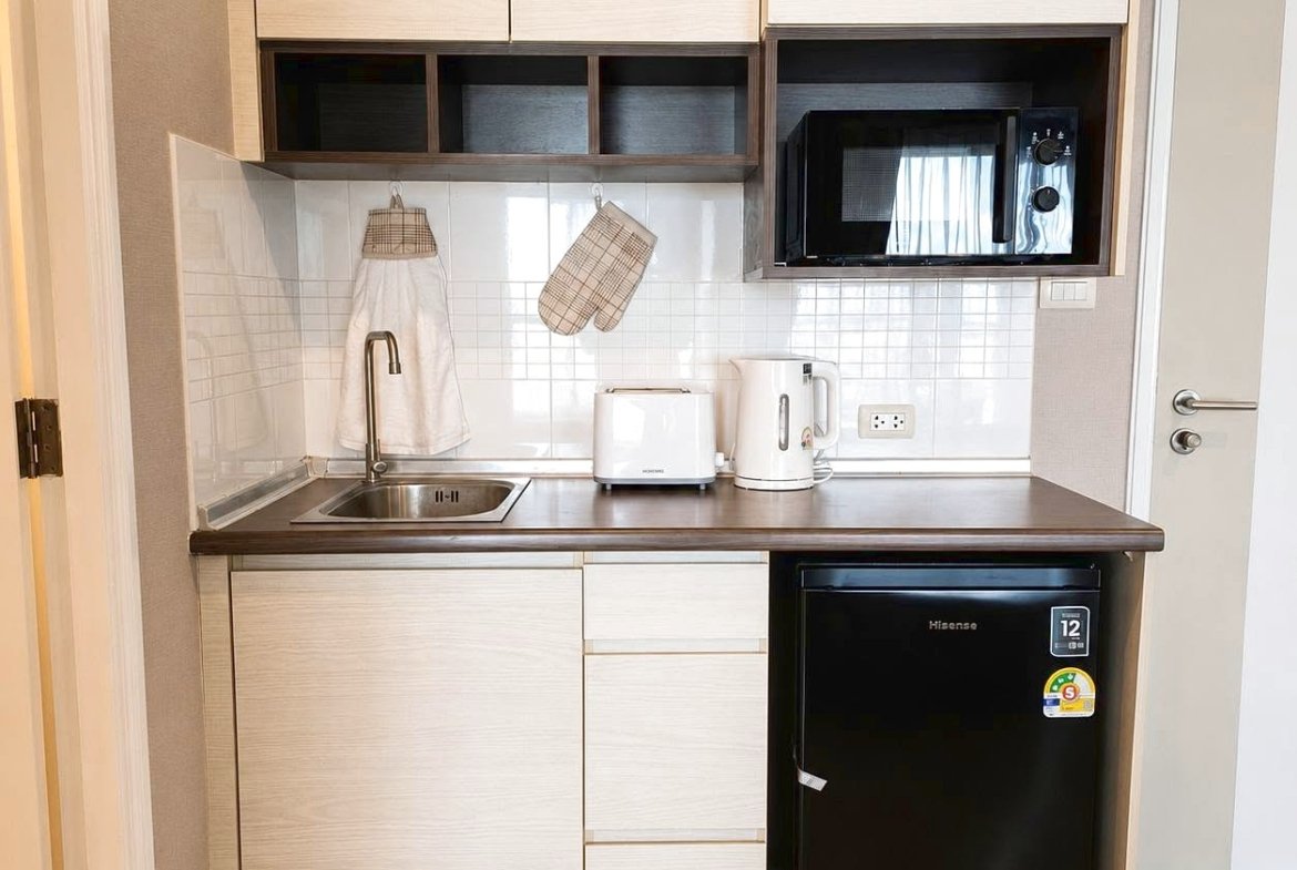 Compact kitchen with a stainless steel sink, light wood cabinets, white tiled backsplash, and small appliances (toaster and electric kettle) on the counter; microwave in a dark wood alcove and a black mini fridge underneath.