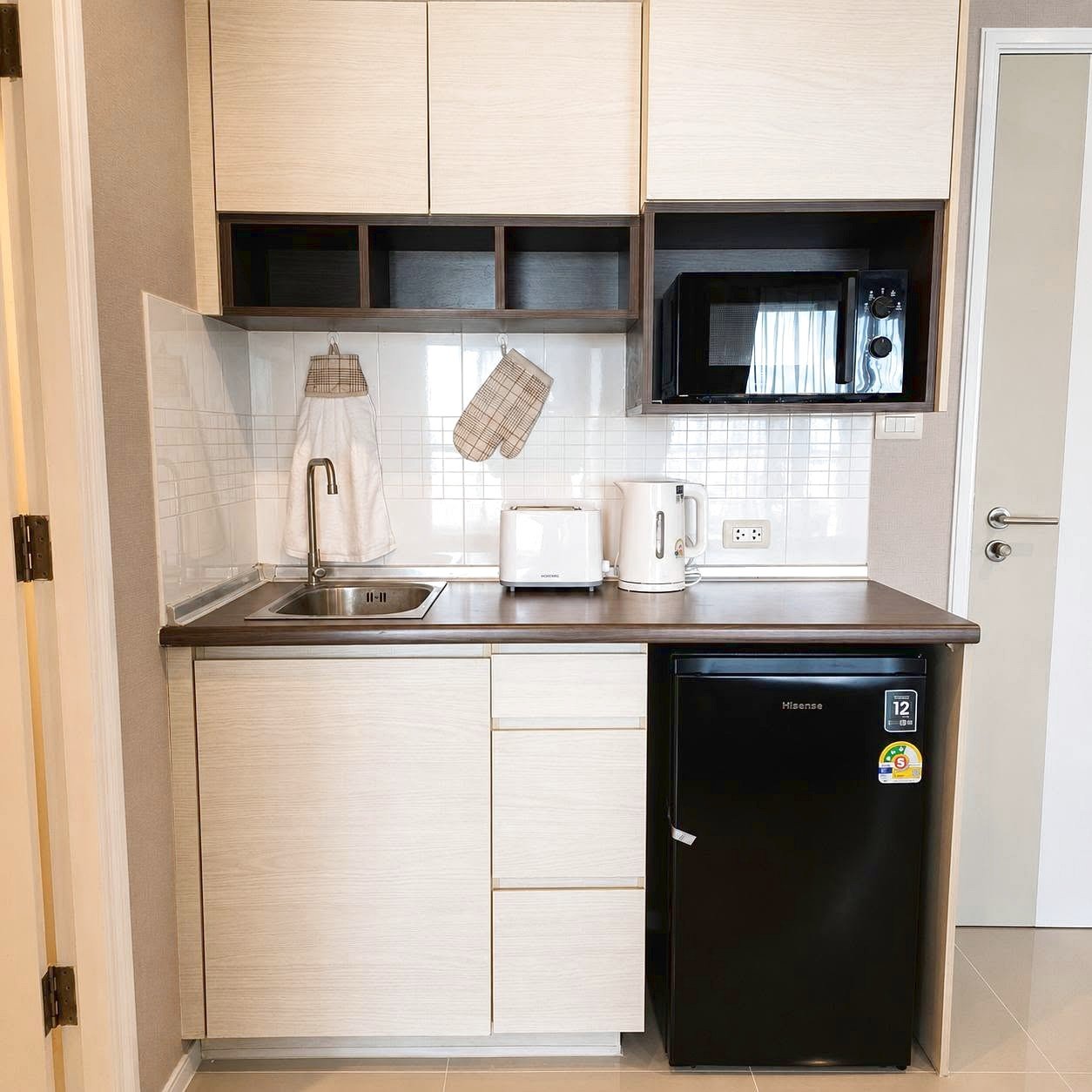 Lumpini_Park_Beach_Sale_Dlc650_14 Compact kitchen with a stainless steel sink, light wood cabinets, white tiled backsplash, and small appliances (toaster and electric kettle) on the counter; microwave in a dark wood alcove and a black mini fridge underneath.
