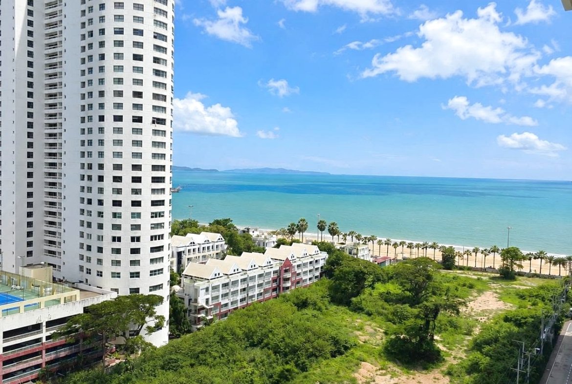 Tall white apartment tower beside a sandy beach and turquoise sea with palm-lined shore.
