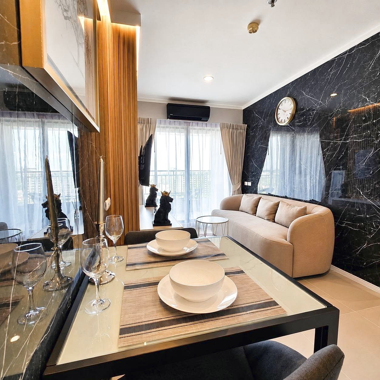Lumpini_Park_Beach_Sale_Dlc650_2 Contemporary living and dining area: glass table set for two with bowls and wine glasses, beige sofa, and a black marble accent wall.