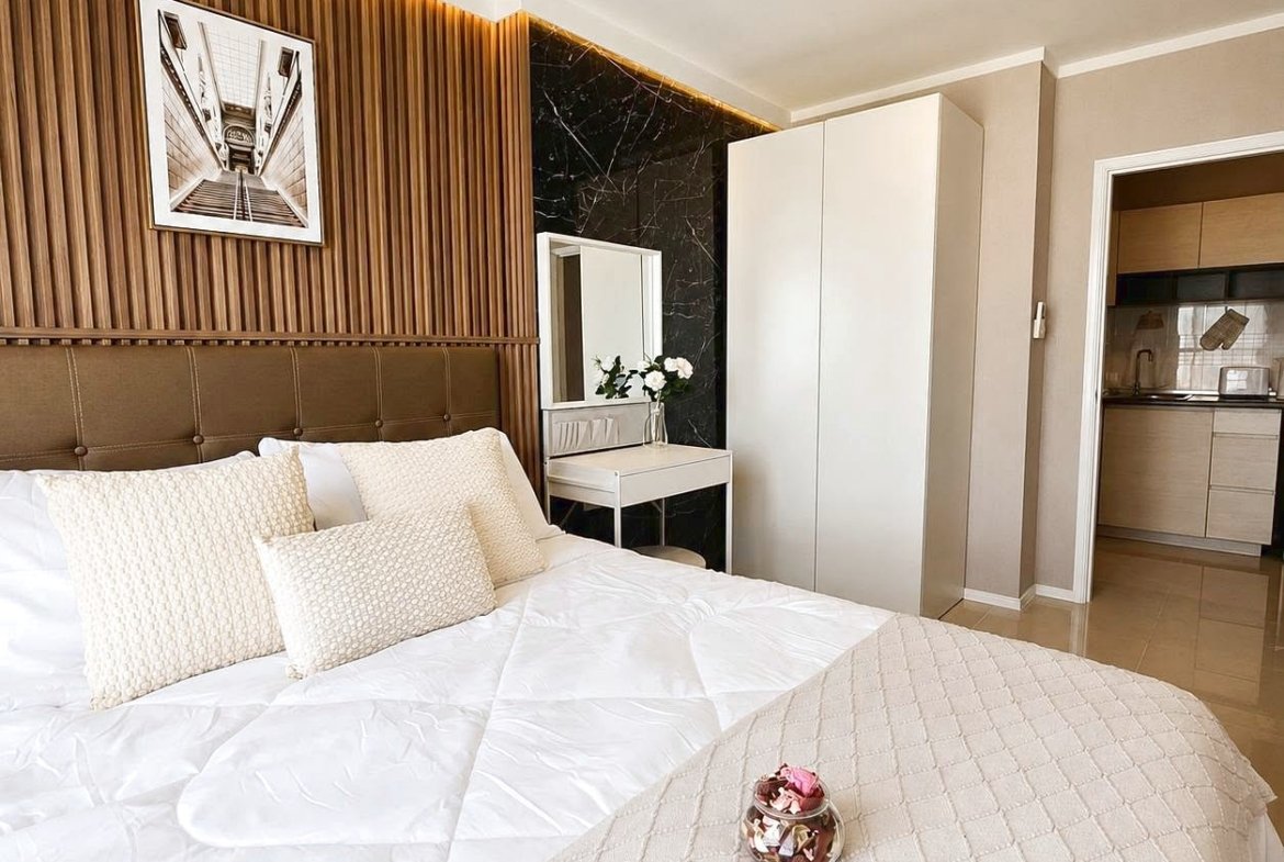 Bedroom with a large white comforter and beige textured pillows against a tufted headboard, wooden slat wall, and framed black-and-white art above the bed.
