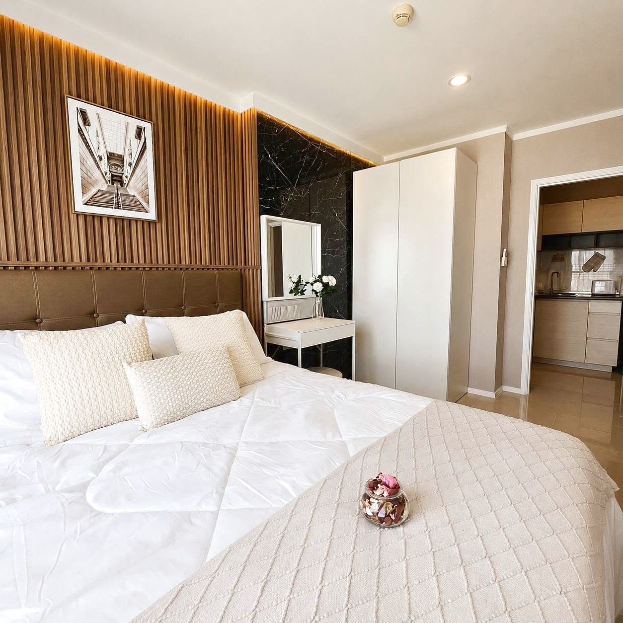 Lumpini_Park_Beach_Sale_Dlc650_3 Bedroom with a large white comforter and beige textured pillows against a tufted headboard, wooden slat wall, and framed black-and-white art above the bed.