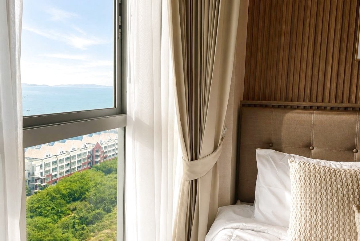 Hotel room with a tufted beige headboard, white bedding, and a large window revealing the ocean and distant buildings outside.