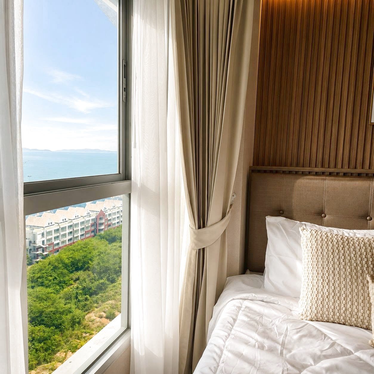 Lumpini_Park_Beach_Sale_Dlc650_5 Hotel room with a tufted beige headboard, white bedding, and a large window revealing the ocean and distant buildings outside.