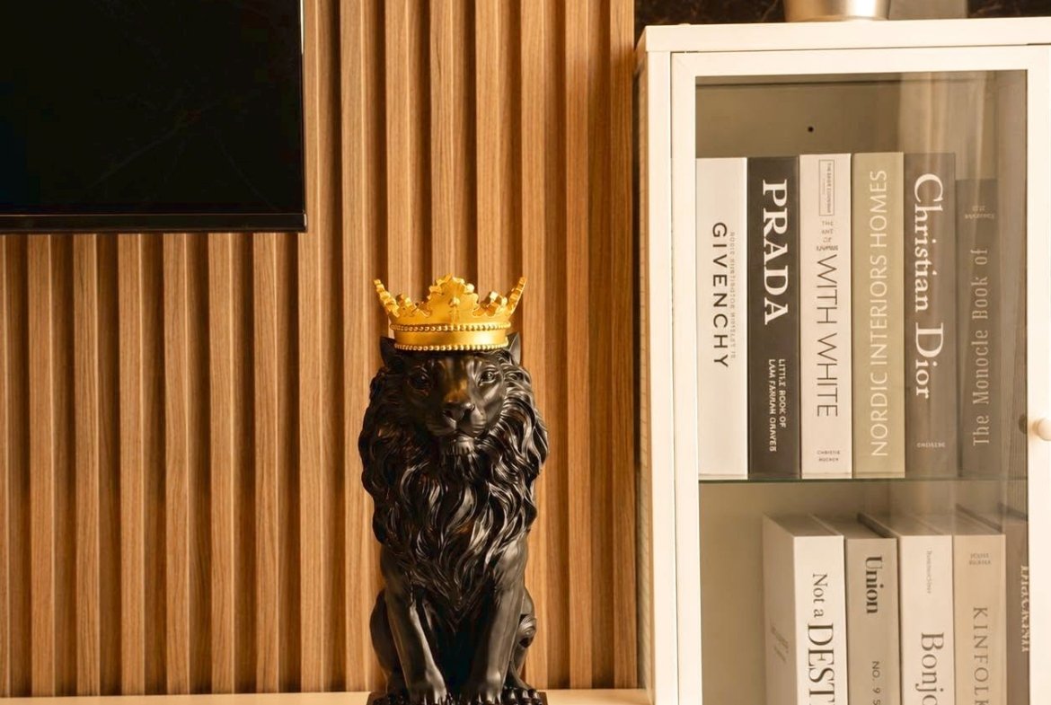 Decorative black lion statue wearing a gold crown on a white shelf beside a glass-front bookcase with neutral books and a plant nearby.