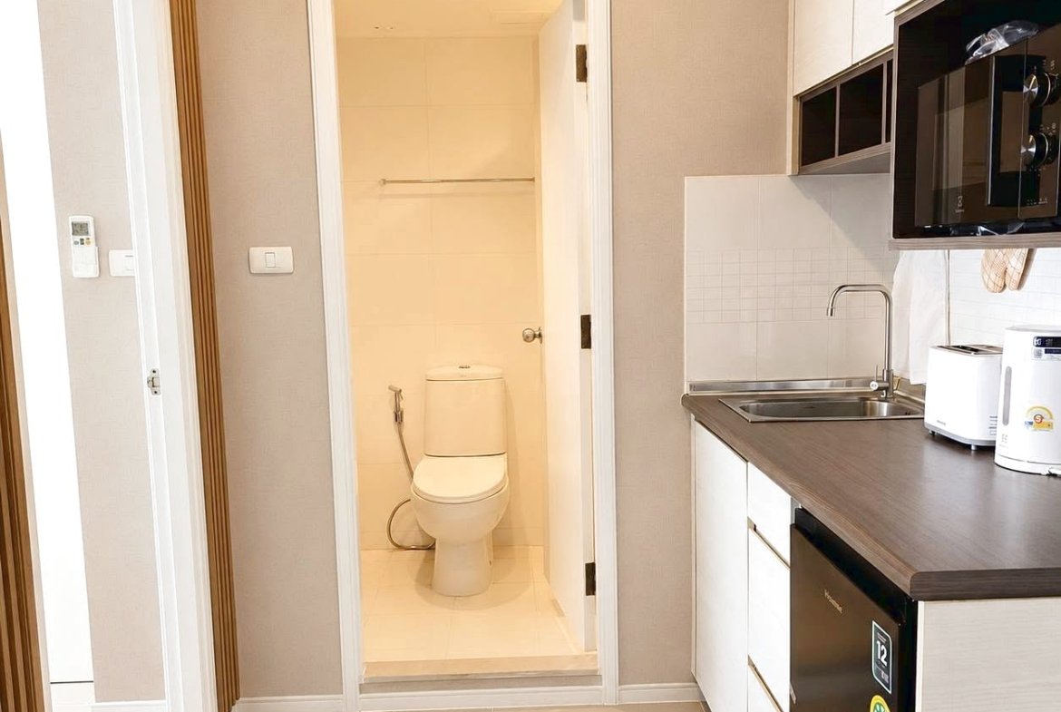 Compact kitchen area with countertop, sink, microwave, kettle, and mini fridge; doorway leads to a small tiled bathroom with a toilet.