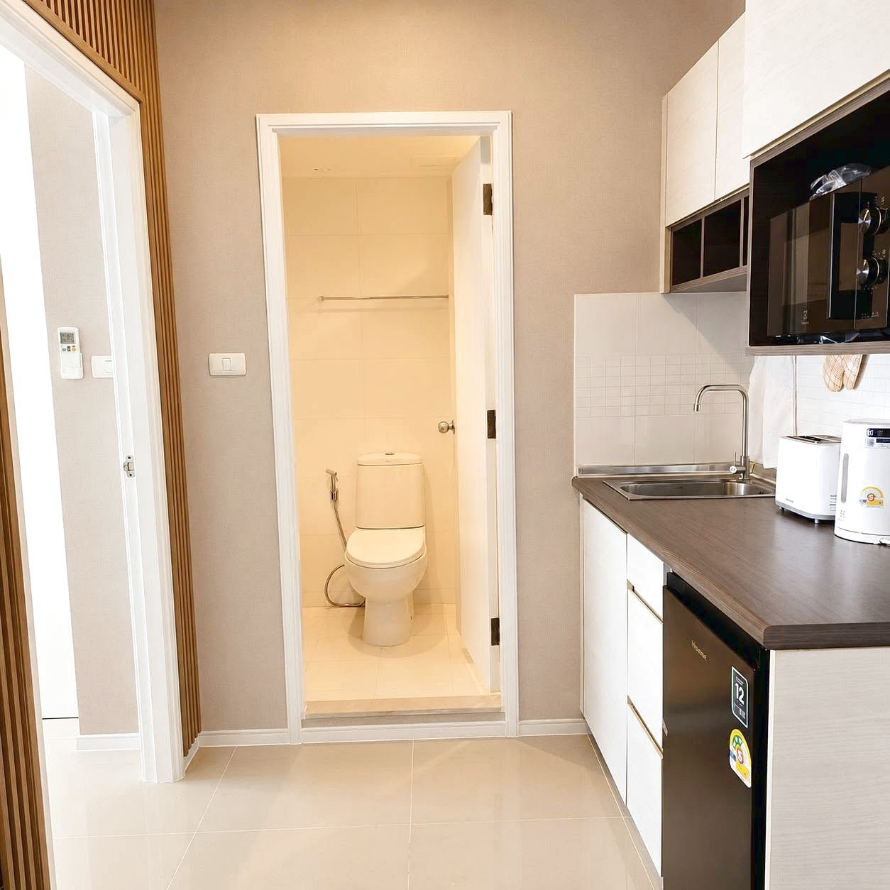 Lumpini_Park_Beach_Sale_Dlc650_9 Compact kitchen area with countertop, sink, microwave, kettle, and mini fridge; doorway leads to a small tiled bathroom with a toilet.