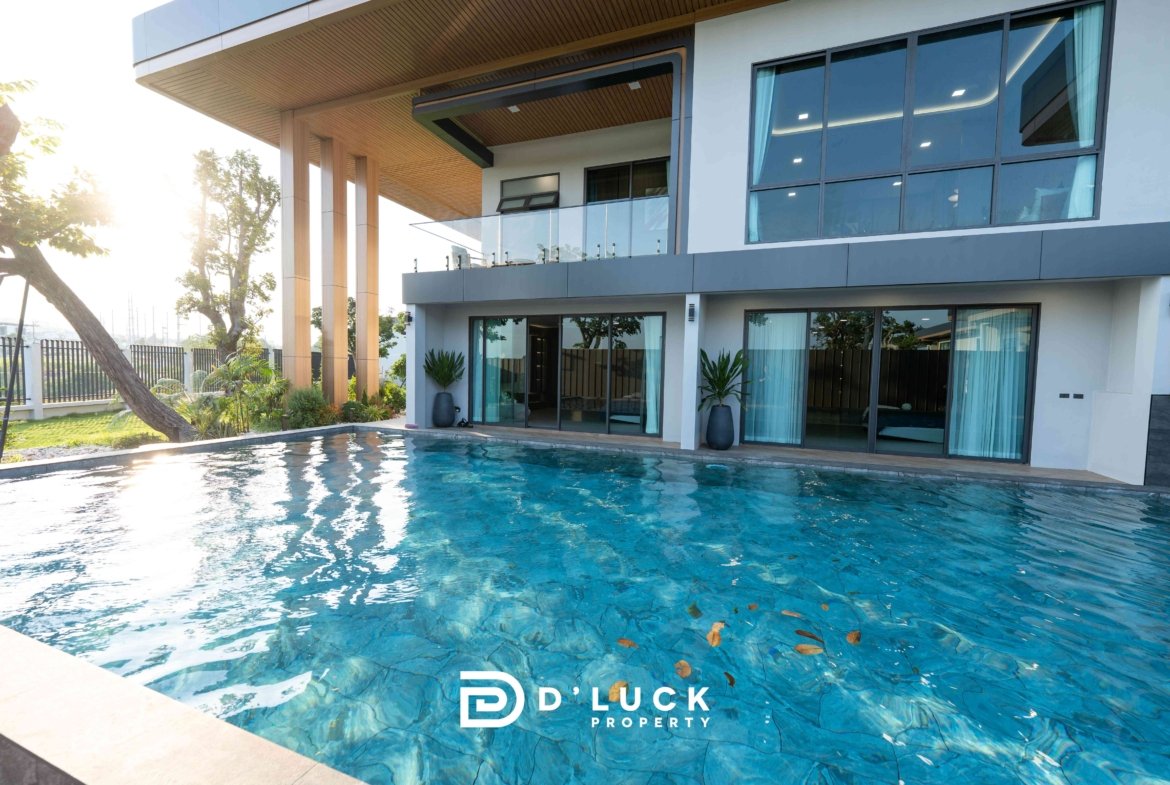 Modern two-story house with glass balcony, large windows, and a blue swimming pool in the foreground; D Luck Property logo visible at bottom.