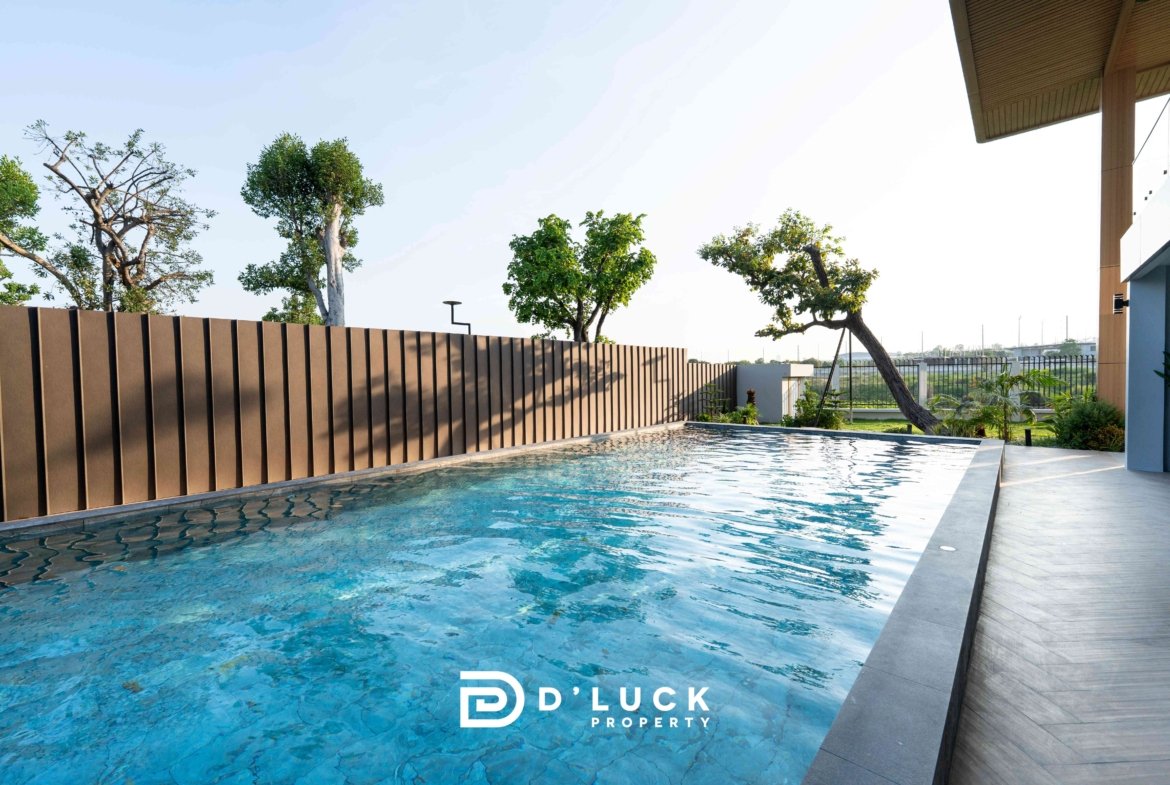 Poolside view of a modern property with blue water, a tall brown slatted fence, and trees along the edge; logo 'D’LUCK PROPERTY' visible on the pool surface