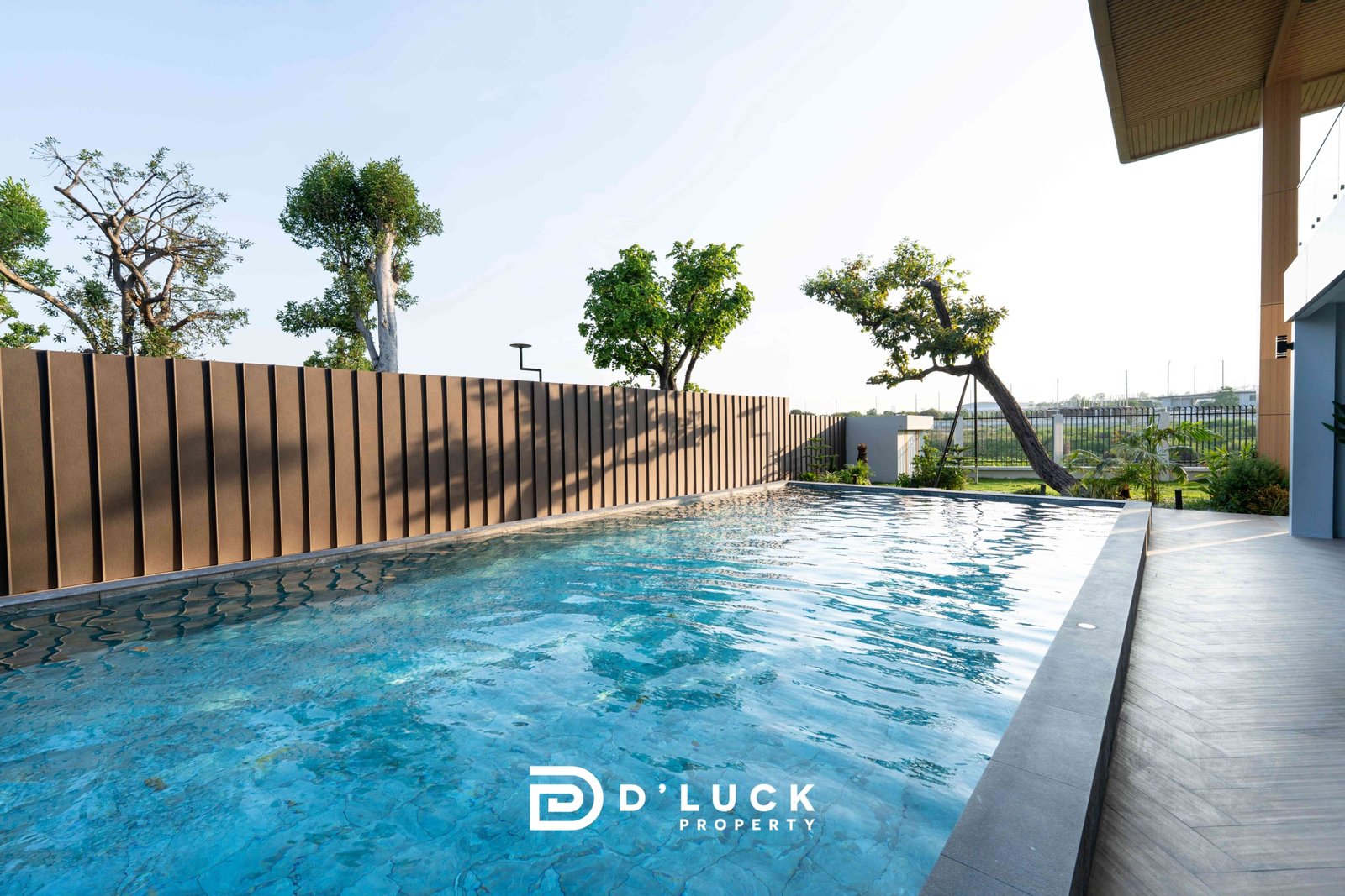 M-Mountian-M3 117 Poolside view of a modern property with blue water, a tall brown slatted fence, and trees along the edge; logo 'D’LUCK PROPERTY' visible on the pool surface