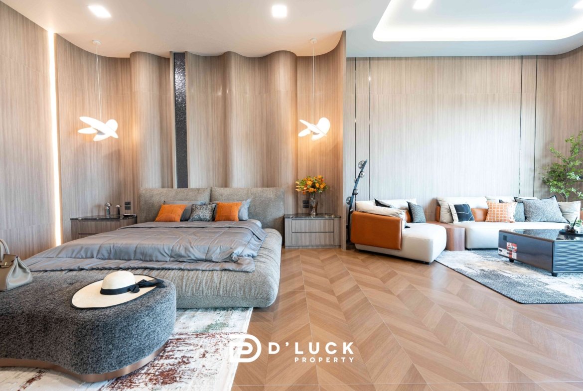 Luxurious hotel suite living area with a gray upholstered bed, orange pillows, a matching gray sofa, and sculptural white pendant lights against wood-paneled walls.