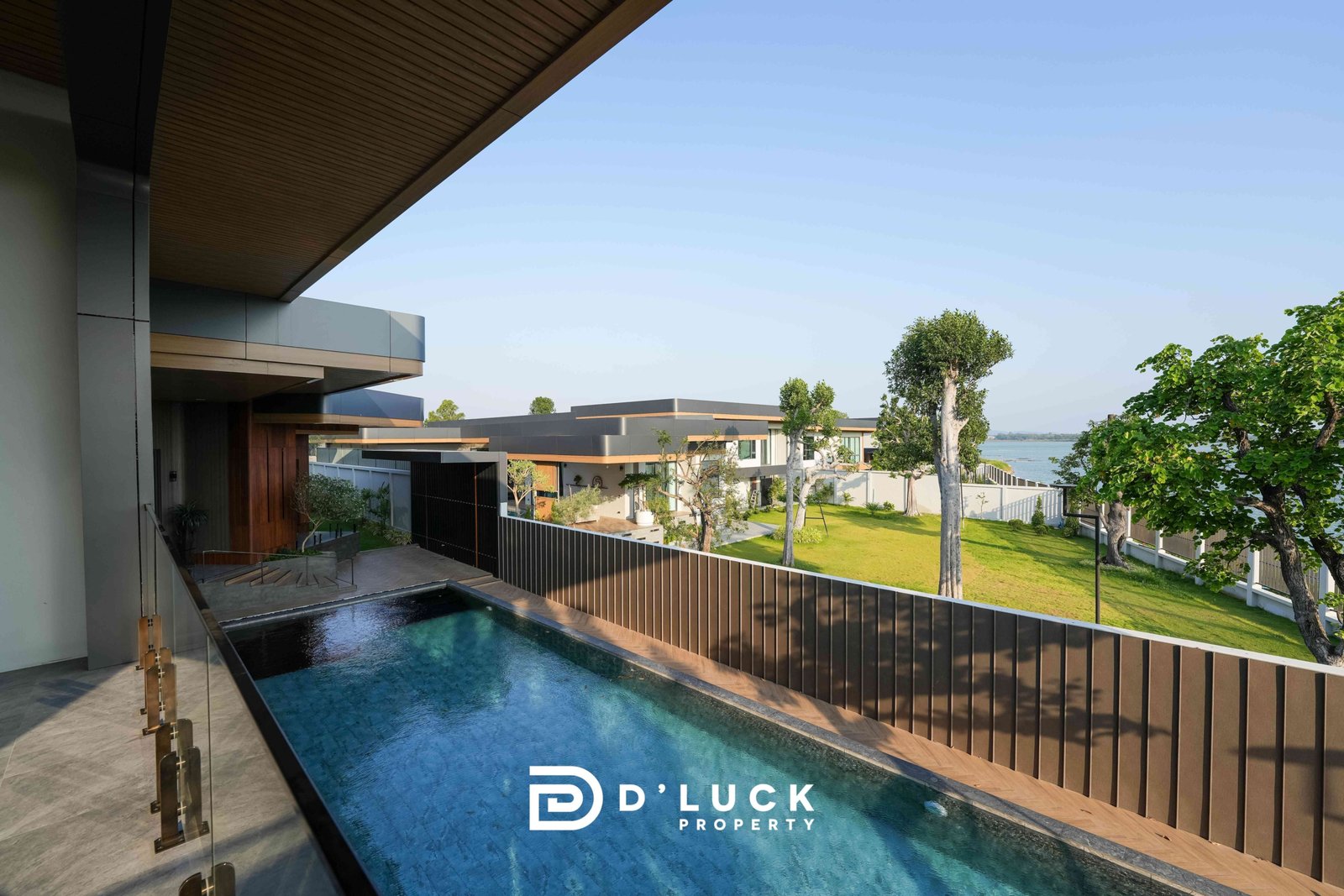 M-Mountian-M3 50 Modern waterfront property with a long rectangular pool, glass railing, and wooden overhang, overlooking green lawns and trees toward the sea with a logo bottom center.
