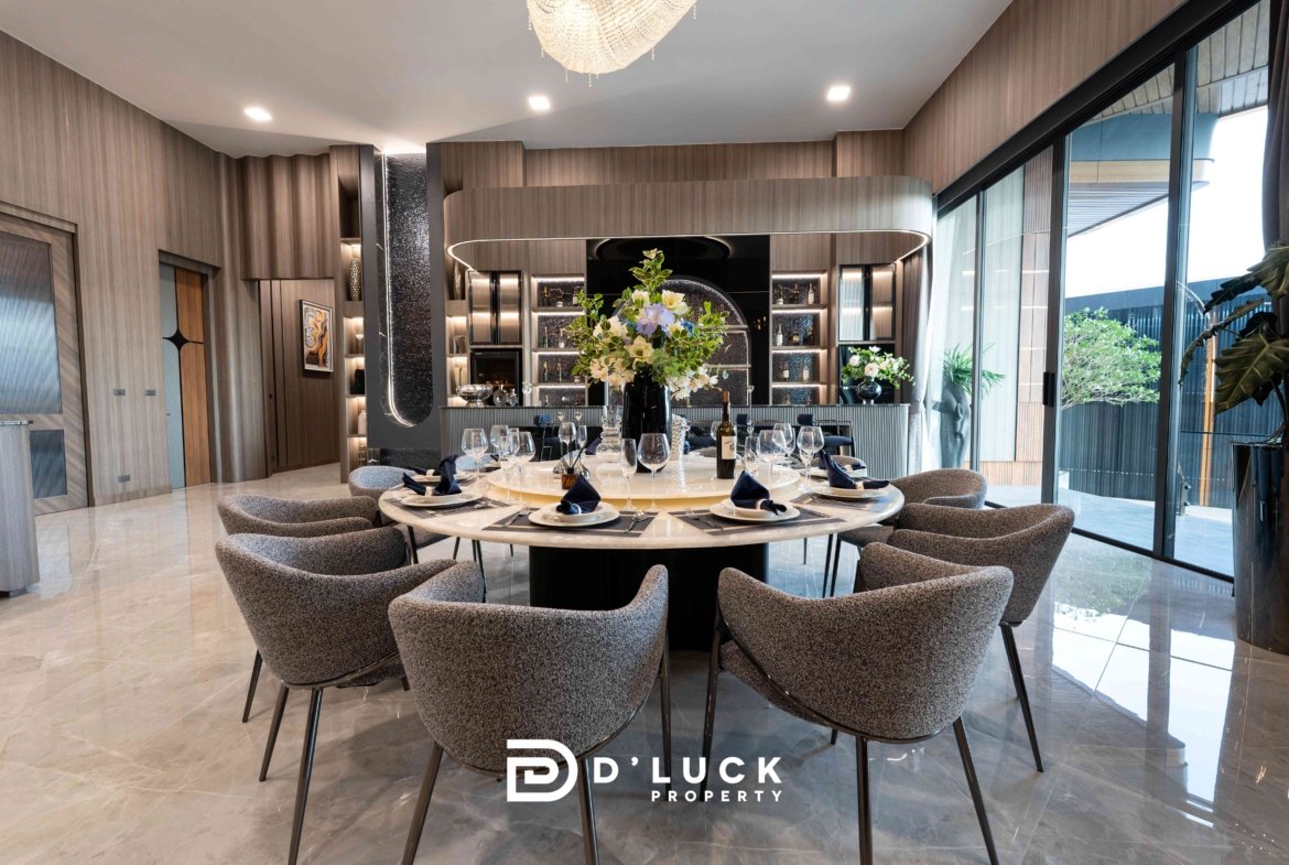 Luxurious dining room with a round table set for eight, a tall floral centerpiece, wine glasses and a curved wood bar against glass walls.