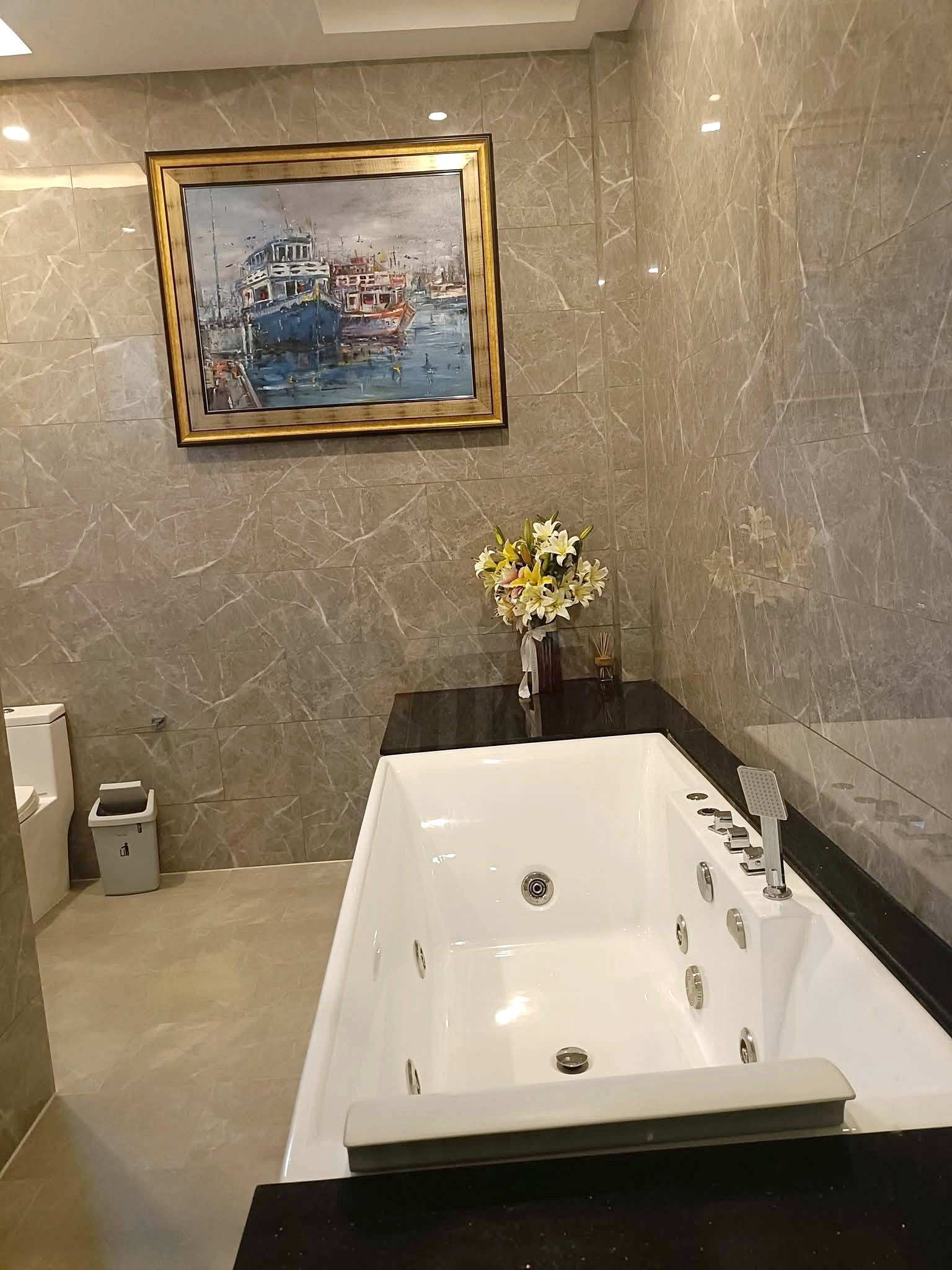 Luxurious bathroom with a white whirlpool tub, black countertop, and a framed maritime painting on marble walls, plus a floral bouquet on the counter.