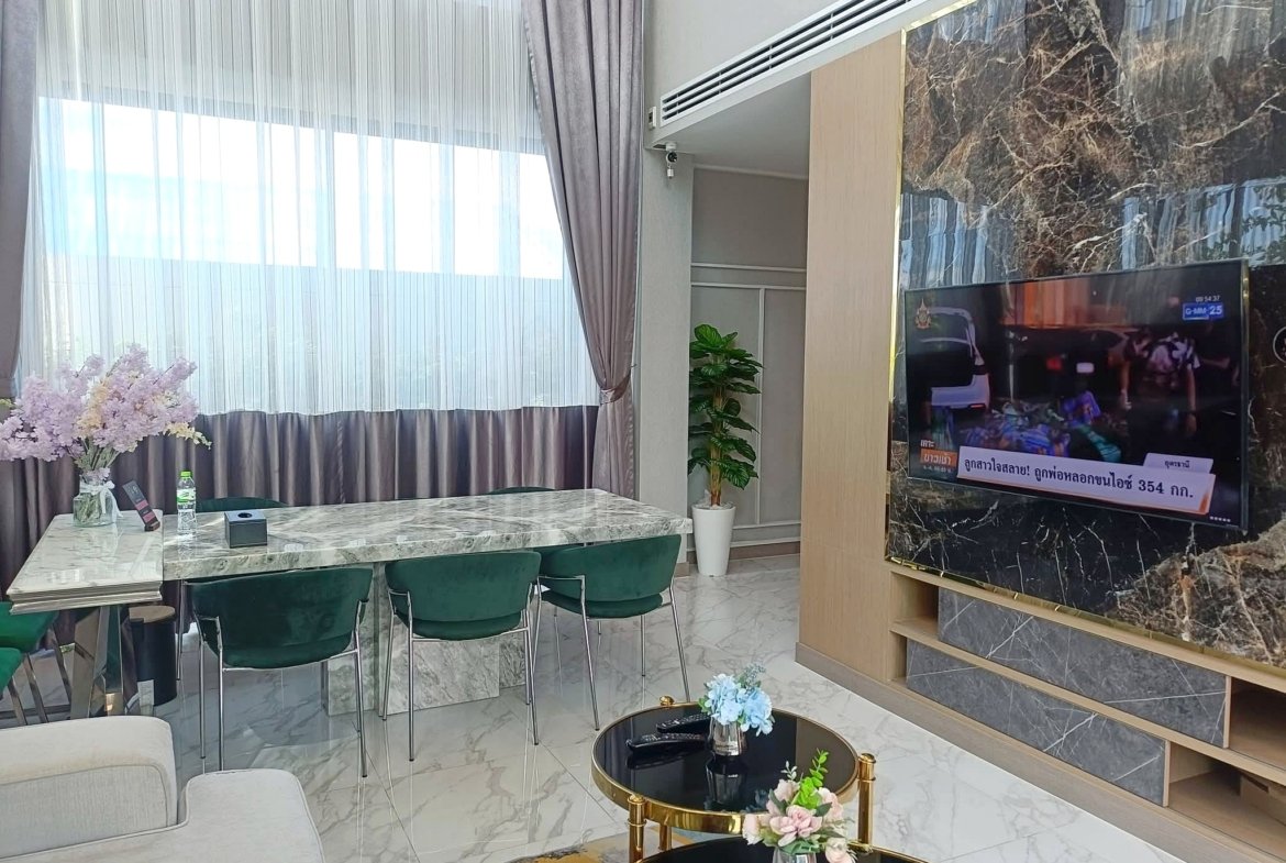 Modern living room with marble floors, glass dining table, green chairs, large windows with sheer curtains, a potted plant, and a wall-mounted TV on a marble wall.