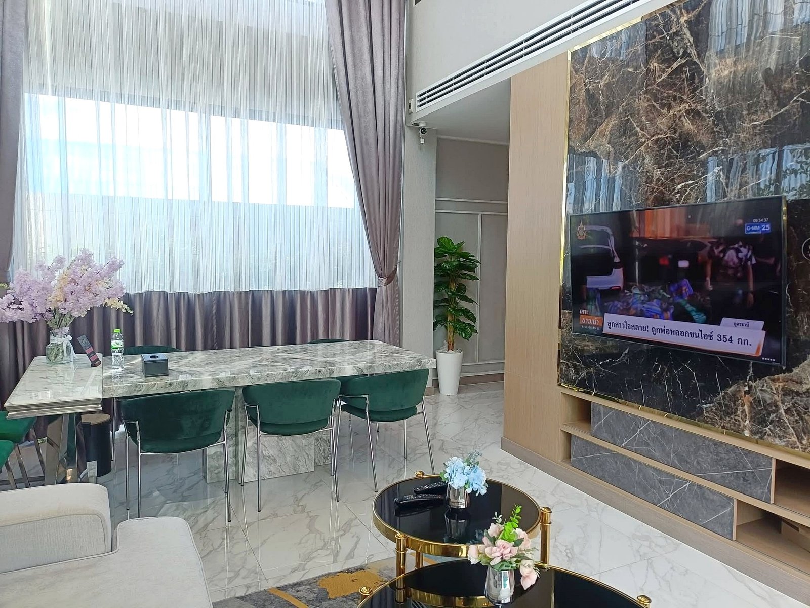 Modern living room with marble floors, glass dining table, green chairs, large windows with sheer curtains, a potted plant, and a wall-mounted TV on a marble wall.