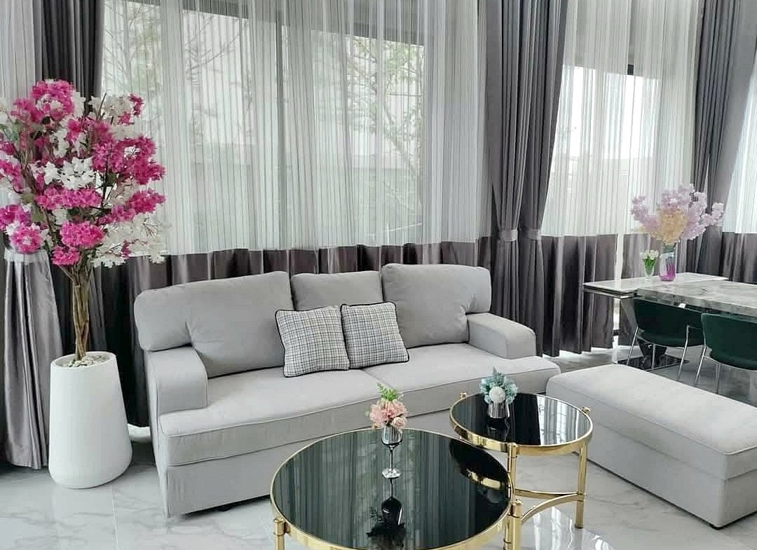 Modern living room with a gray sofa, matching ottoman, glass-top coffee tables with gold frames, and pink blossom arrangements near sheer curtains.