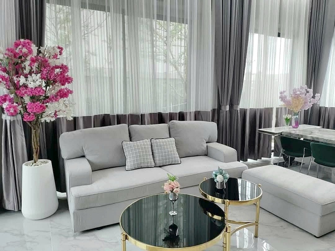 Modern living room with a gray sofa, matching ottoman, glass-top coffee tables with gold frames, and pink blossom arrangements near sheer curtains.