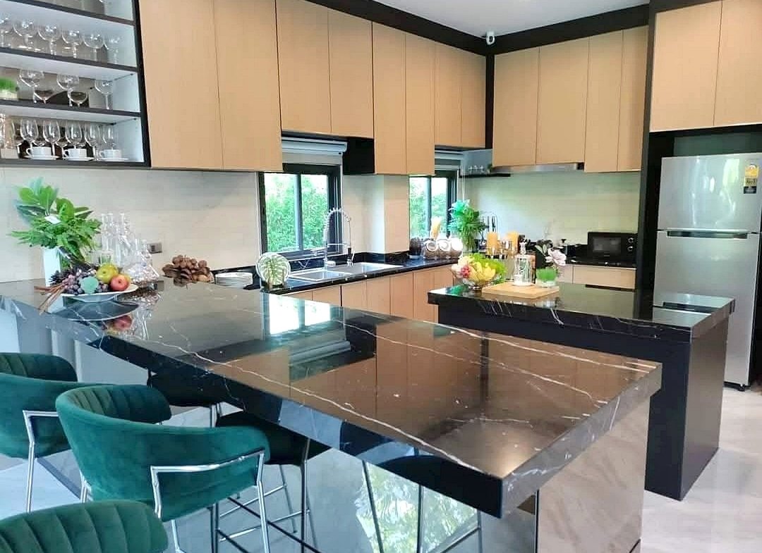 Modern kitchen with a large black marble island, green bar stools, light wood cabinets, and a stainless steel fridge.