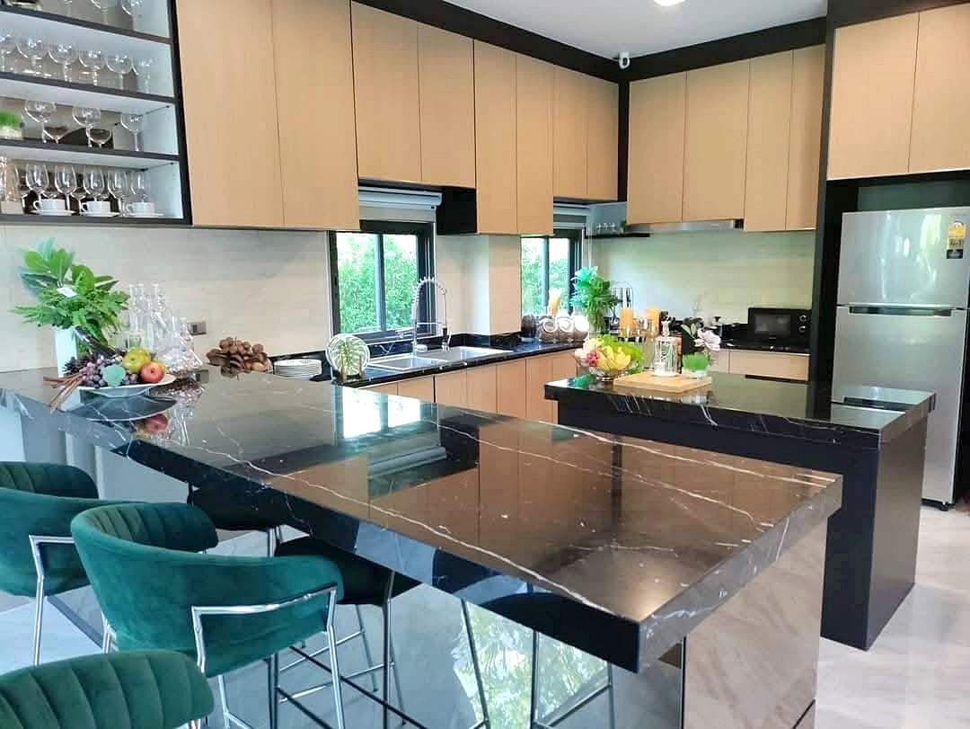 Modern kitchen with a large black marble island, green bar stools, light wood cabinets, and a stainless steel fridge.