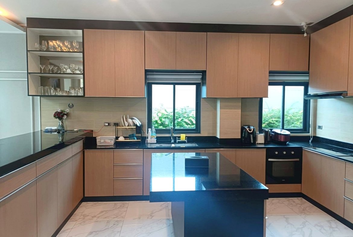 Modern kitchen with light wooden cabinets, black countertops, and an island; glassware on open shelves to the left.