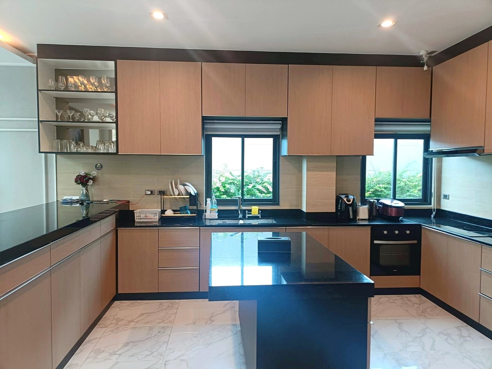 Modern kitchen with light wooden cabinets, black countertops, and an island; glassware on open shelves to the left.
