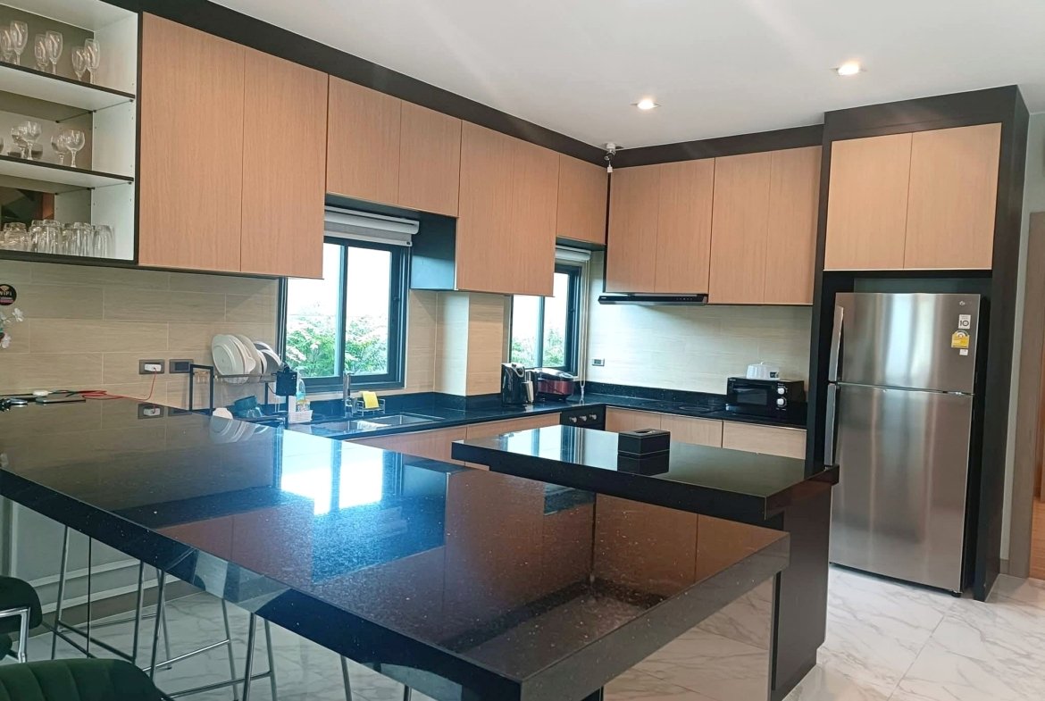 Modern kitchen with light wood cabinets, black granite island, and stainless steel fridge; glass-front shelves on the left and large windows provide natural light.