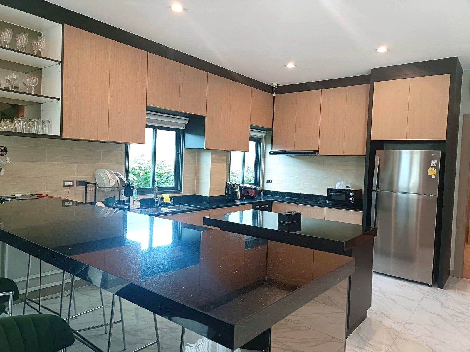 Modern kitchen with light wood cabinets, black granite island, and stainless steel fridge; glass-front shelves on the left and large windows provide natural light.