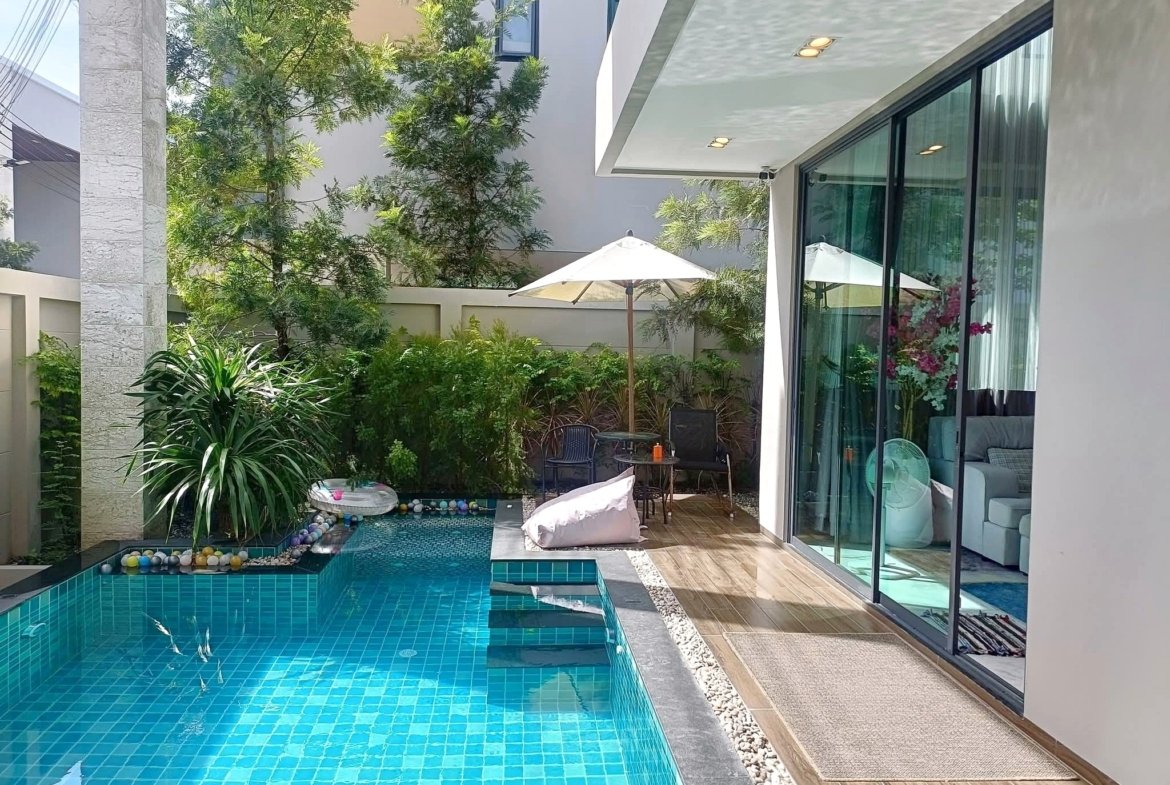 Outdoor courtyard with a turquoise tiled pool beside a wooden deck, glass sliding doors, and lush greenery including tall trees and shrubs under a shaded patio with a white umbrella.