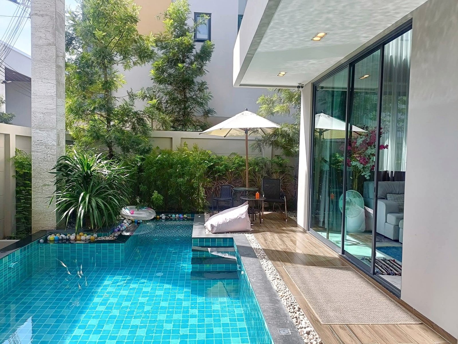 Outdoor courtyard with a turquoise tiled pool beside a wooden deck, glass sliding doors, and lush greenery including tall trees and shrubs under a shaded patio with a white umbrella.