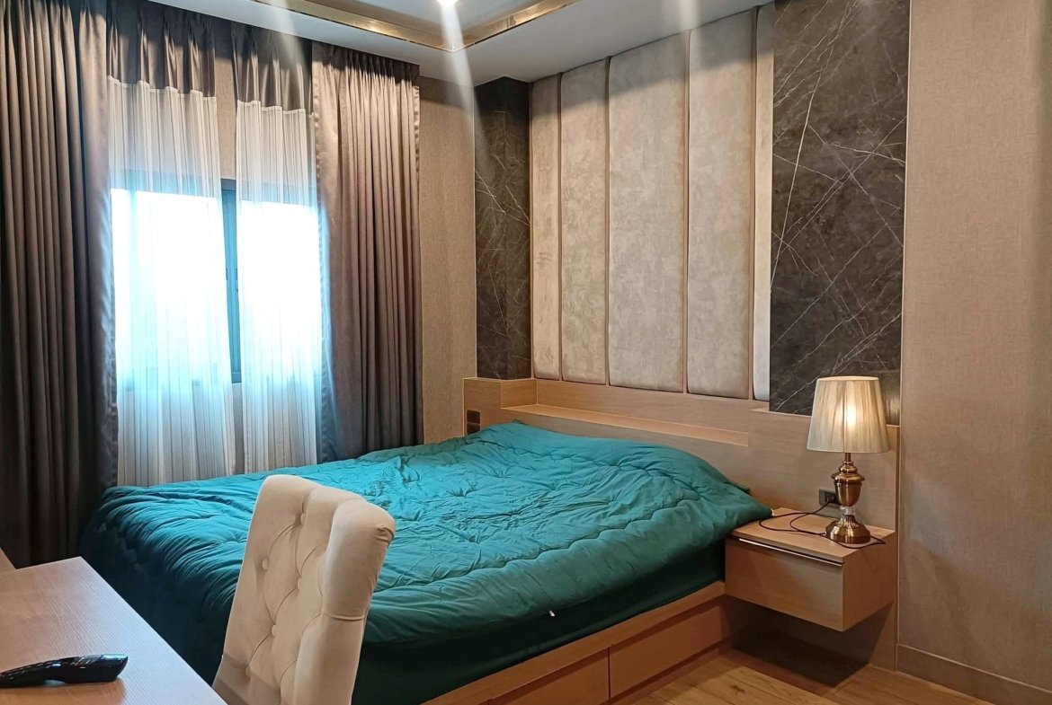 Teal quilted bed in a modern bedroom with beige padded wall panels, a wooden nightstand, and a lamp beside the bed.