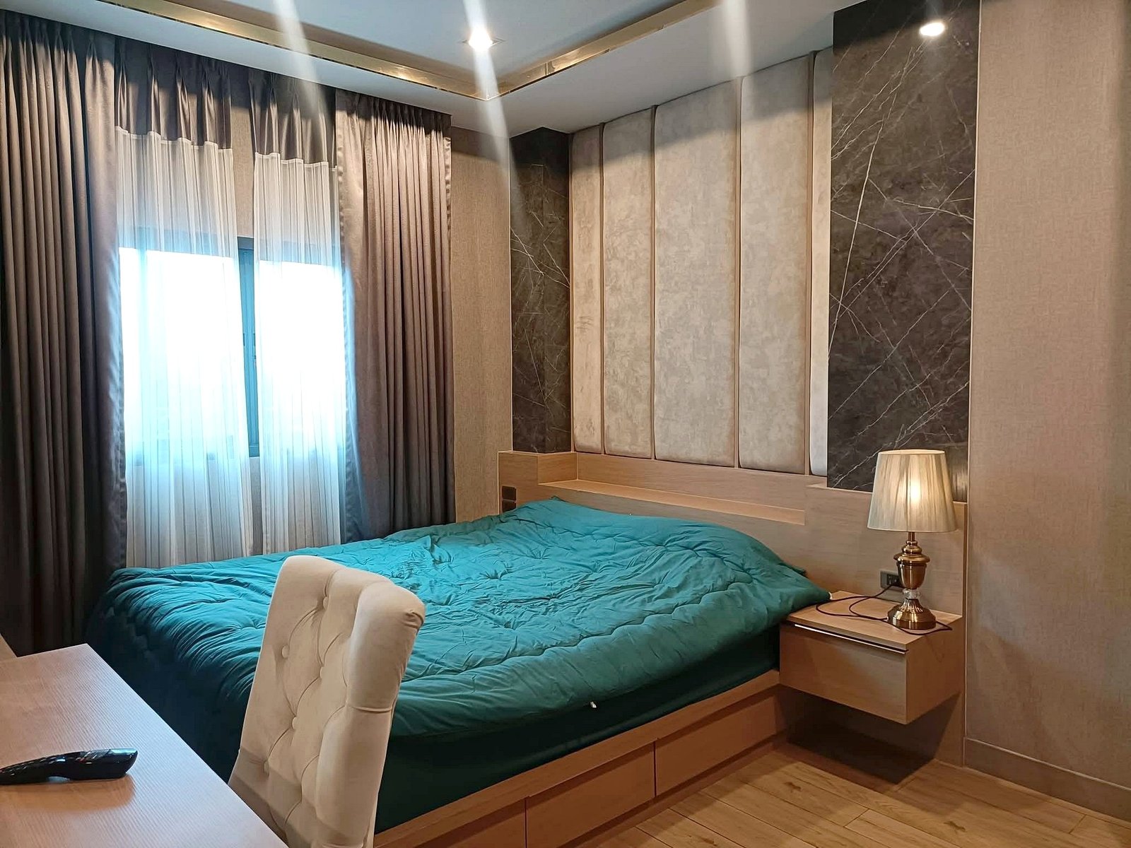 Teal quilted bed in a modern bedroom with beige padded wall panels, a wooden nightstand, and a lamp beside the bed.