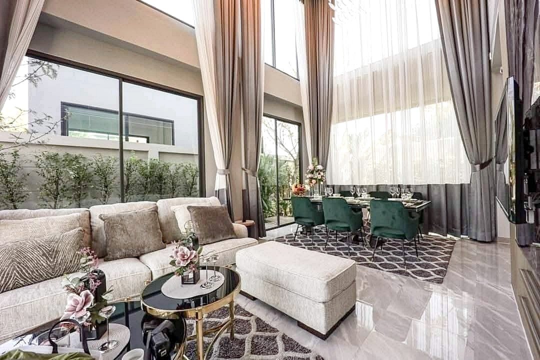 Bright open-plan living and dining area with a beige sectional, glass coffee tables, and a dining set of teal chairs under tall sheer curtains?
