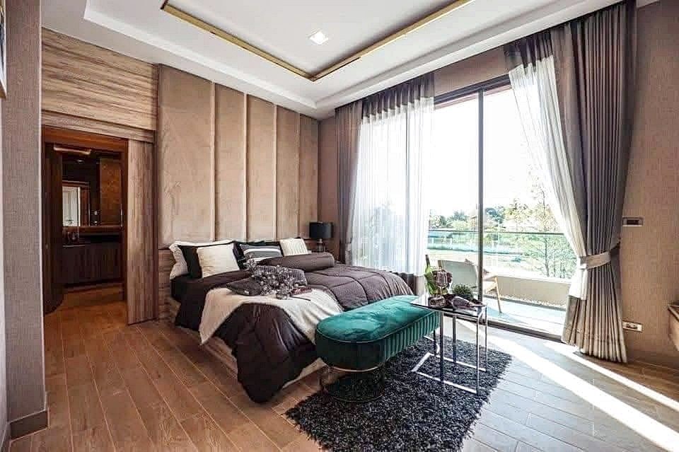 Luxurious bedroom with a king bed, beige padded headboard, teal ottoman, and balcony doors with outdoor seating beyond the glass walls.