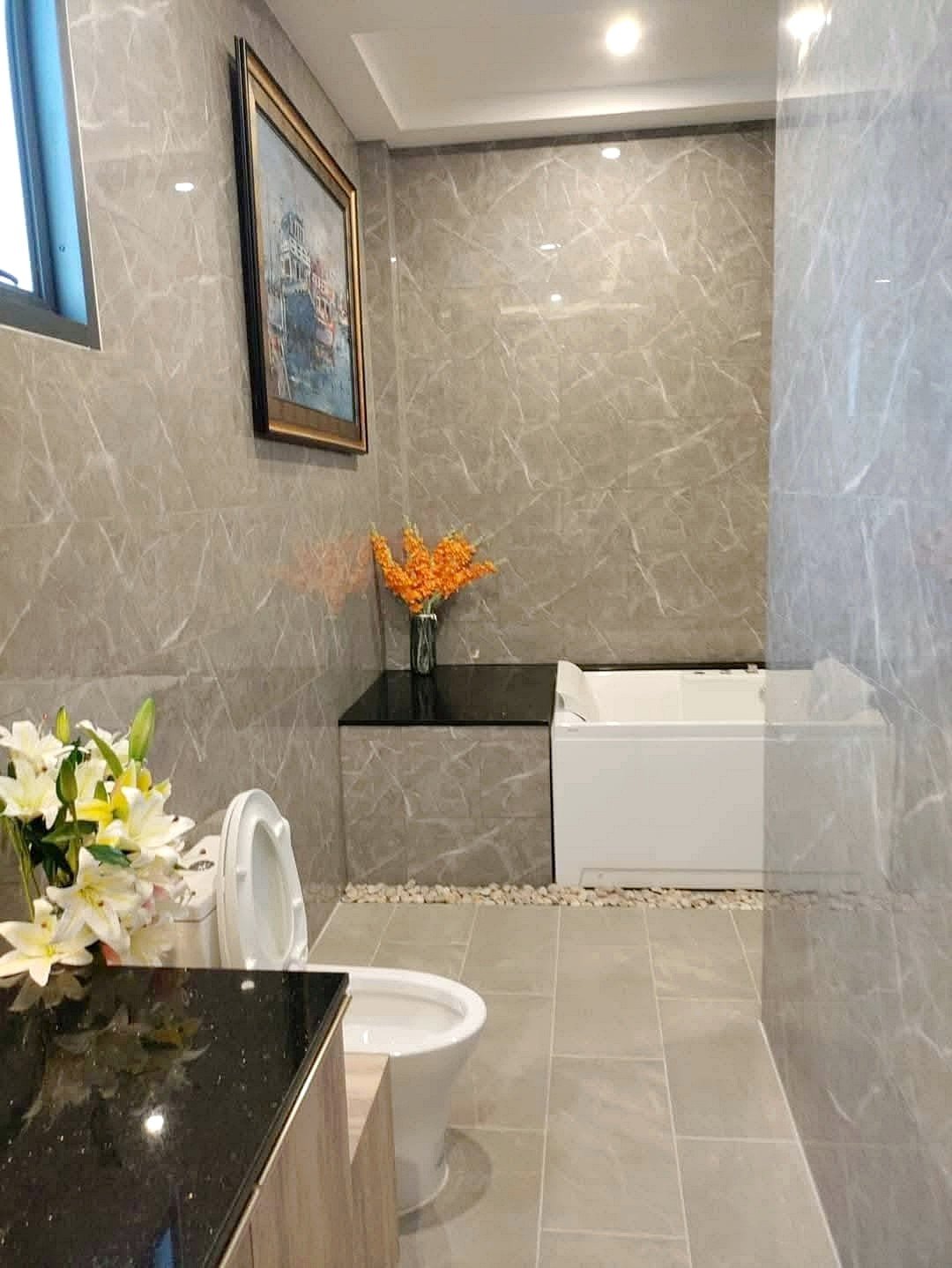 Bathroom with gray marble walls and floor, white bathtub along back wall, black countertop with orange flowers, and a framed painting above.