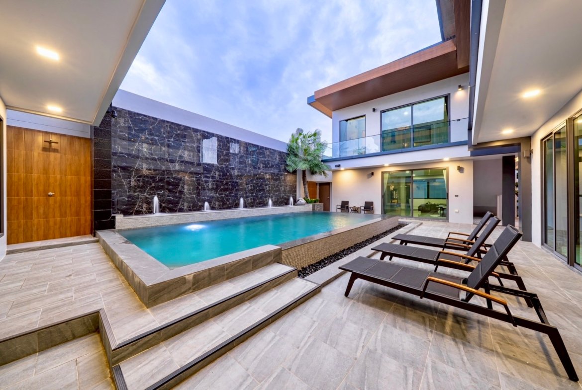 Modern villa courtyard with a rectangular pool, waterfall wall, and lounge chairs on a tiled deck.
