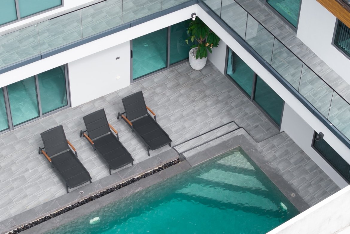 Overhead view of a modern courtyard with a rectangular pool, four black lounge chairs, and gray tiled deck next to glass railings.