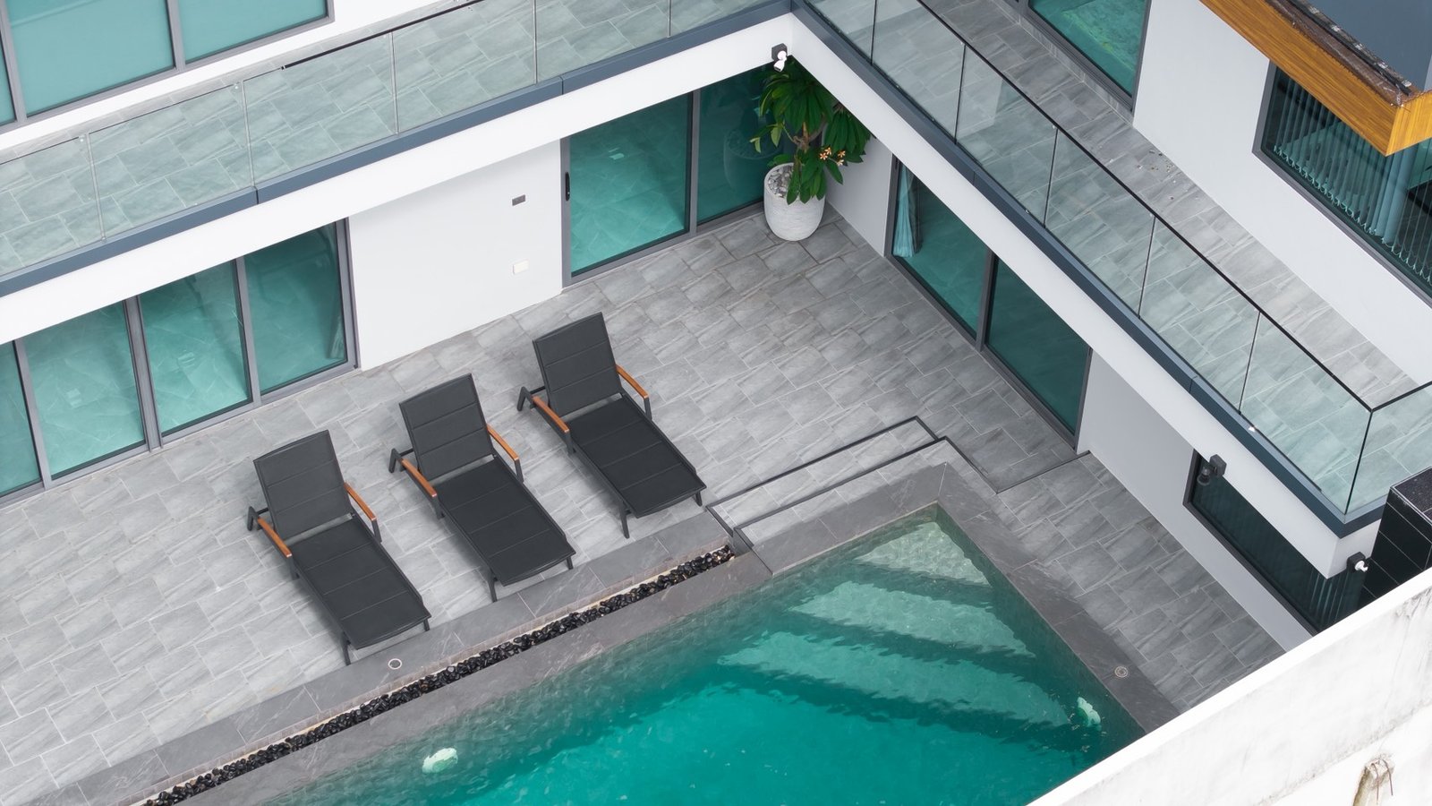 M_Mountain_Dlh518 2 Overhead view of a modern courtyard with a rectangular pool, four black lounge chairs, and gray tiled deck next to glass railings.