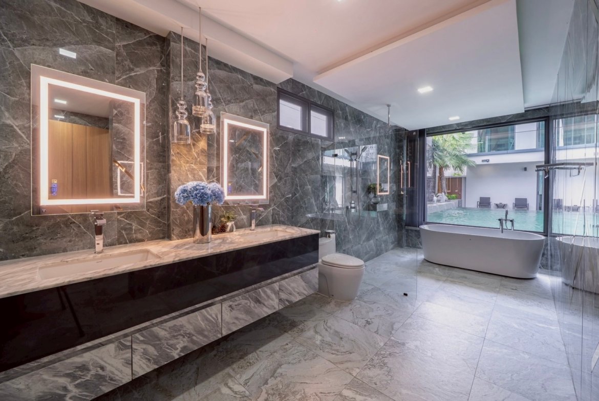 Luxurious marble bathroom with a double-sink vanity, backlit square mirrors, and a freestanding bathtub by a large window overlooking an outdoor pool.